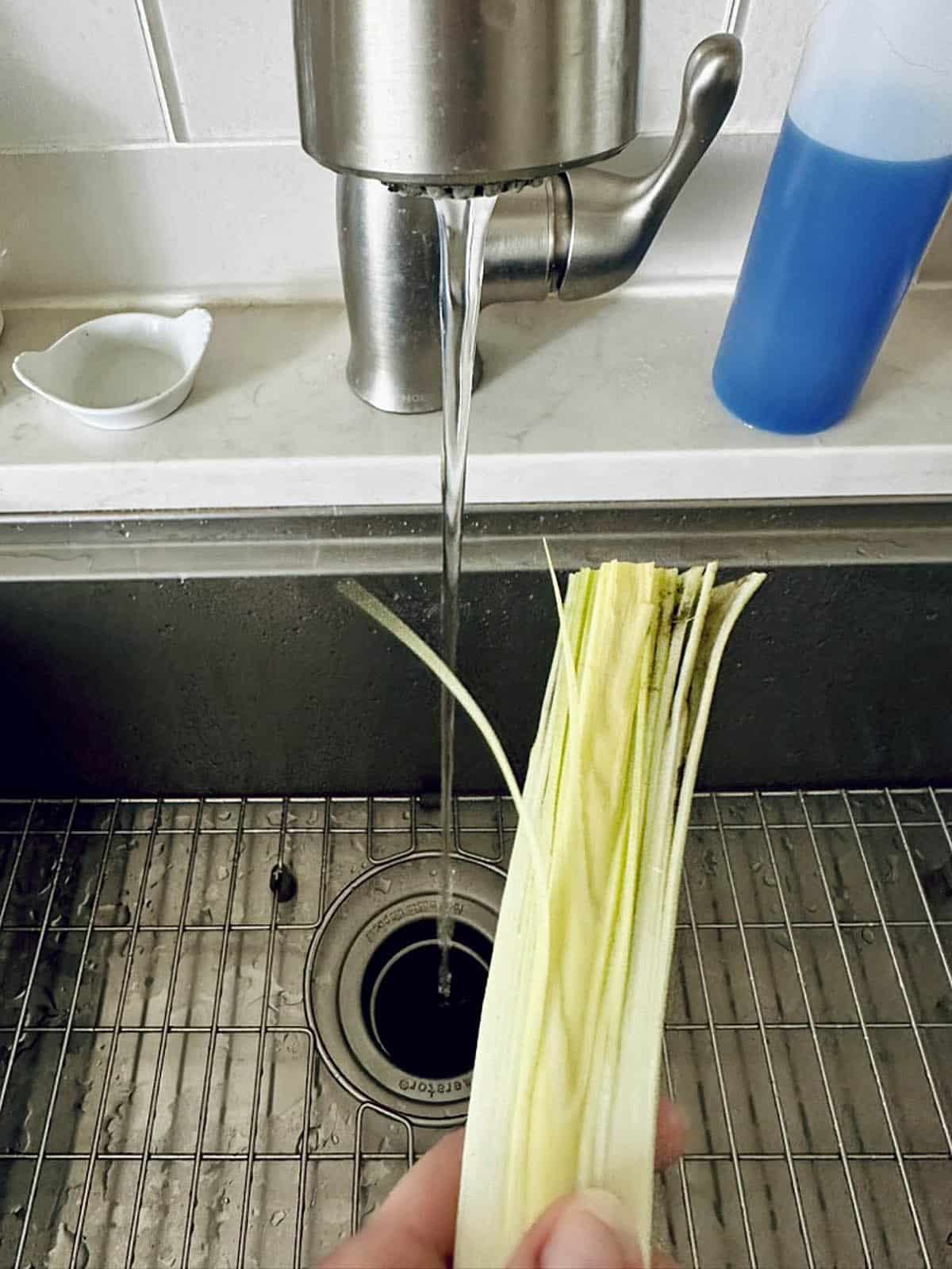 A hand holds a piece of chopped lemongrass under running water at a kitchen sink, preparing fresh ingredients for a comforting Potato Leek Soup. A blue bottle and white dish sit near the faucet.