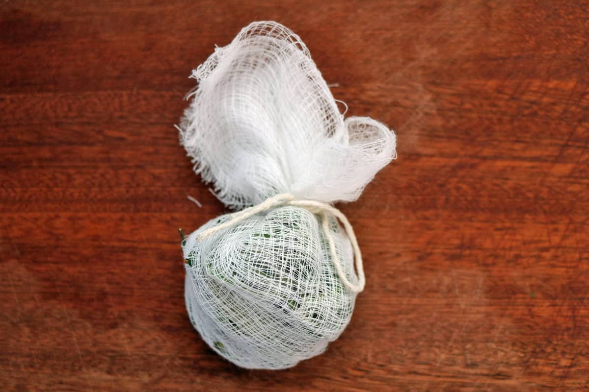 A bundle of herbs wrapped in white cheesecloth and tied with string sits on a wooden surface, perfectly illustrating how to make a bouquet garni and sachet for adding delicate flavor to soups and stews.
