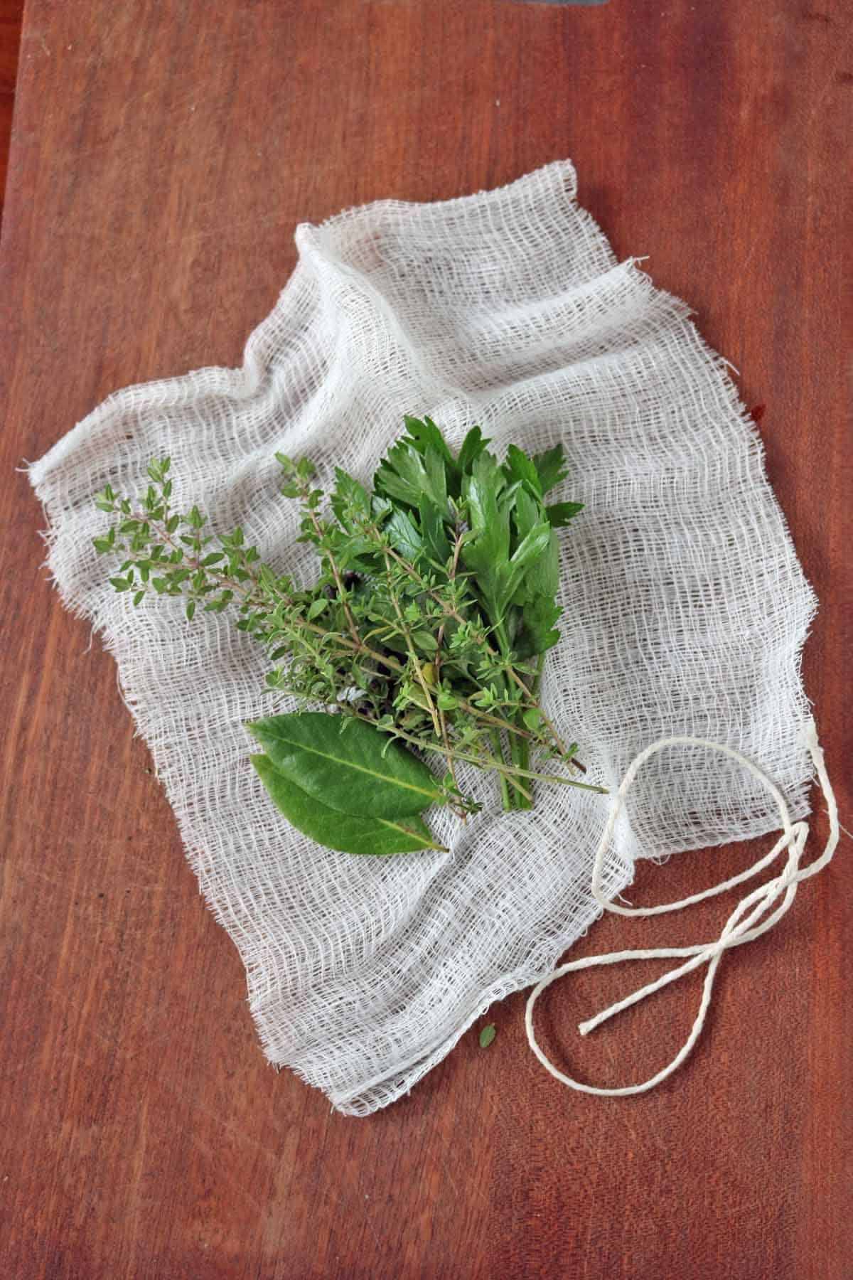 A bundle of fresh herbs on a piece of white cheesecloth with a string, placed on a wooden surface—showing How to Make a bouquet Garni and Sachet for infusing rich flavors into your dishes.