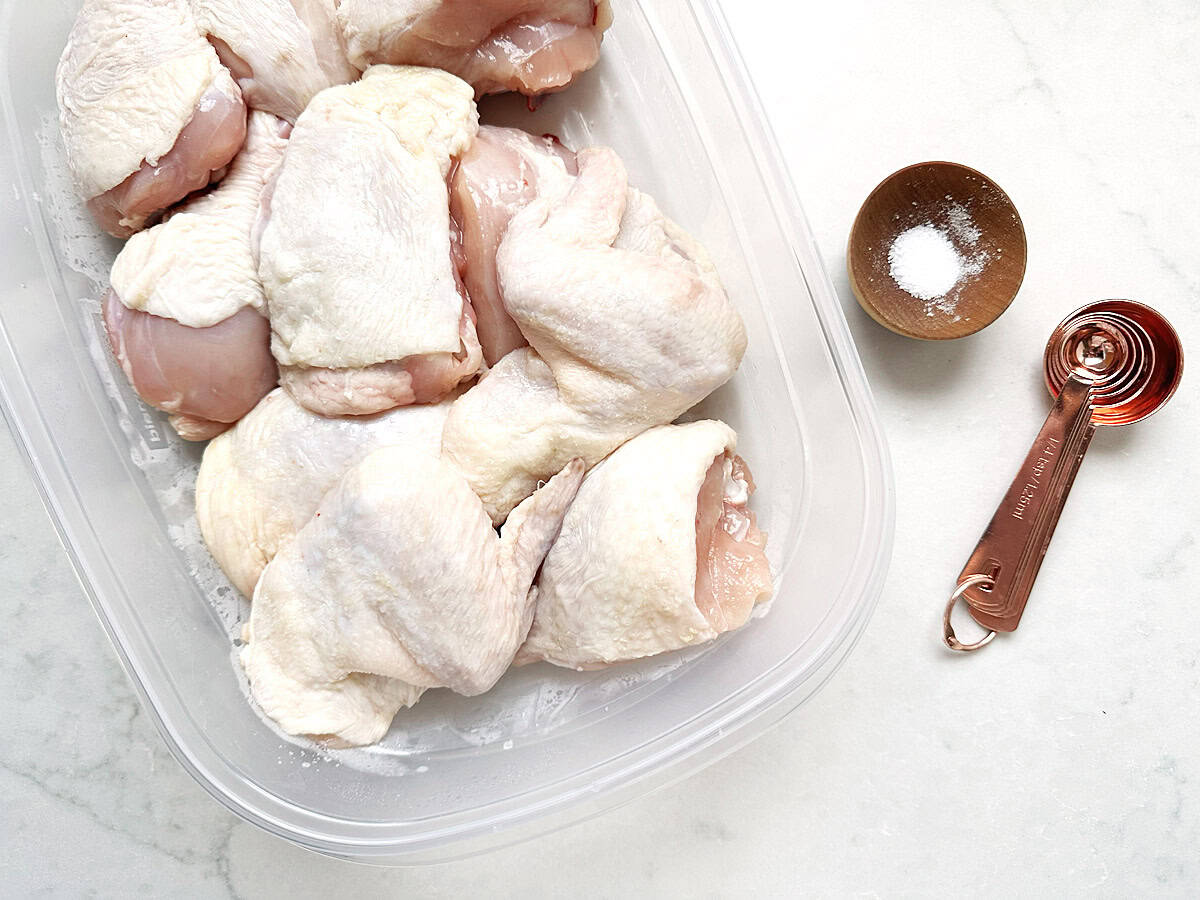 Raw chicken pieces in a clear plastic container are next to a small wooden bowl of salt and a set of copper measuring spoons on a white countertop, ready to be transformed into Foolproof French Grilled Chicken.