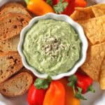 A bowl of green dip topped with shredded cheese and pepper, surrounded by sliced bread, tortilla chips, mini bell peppers, and cilantro on a white plate.