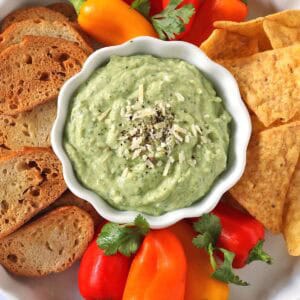 A bowl of green dip topped with shredded cheese and pepper, surrounded by sliced bread, tortilla chips, mini bell peppers, and cilantro on a white plate.