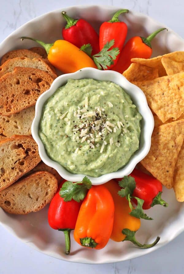 A bowl of green dip topped with shredded cheese and pepper, surrounded by sliced bread, tortilla chips, mini bell peppers, and cilantro on a white plate.