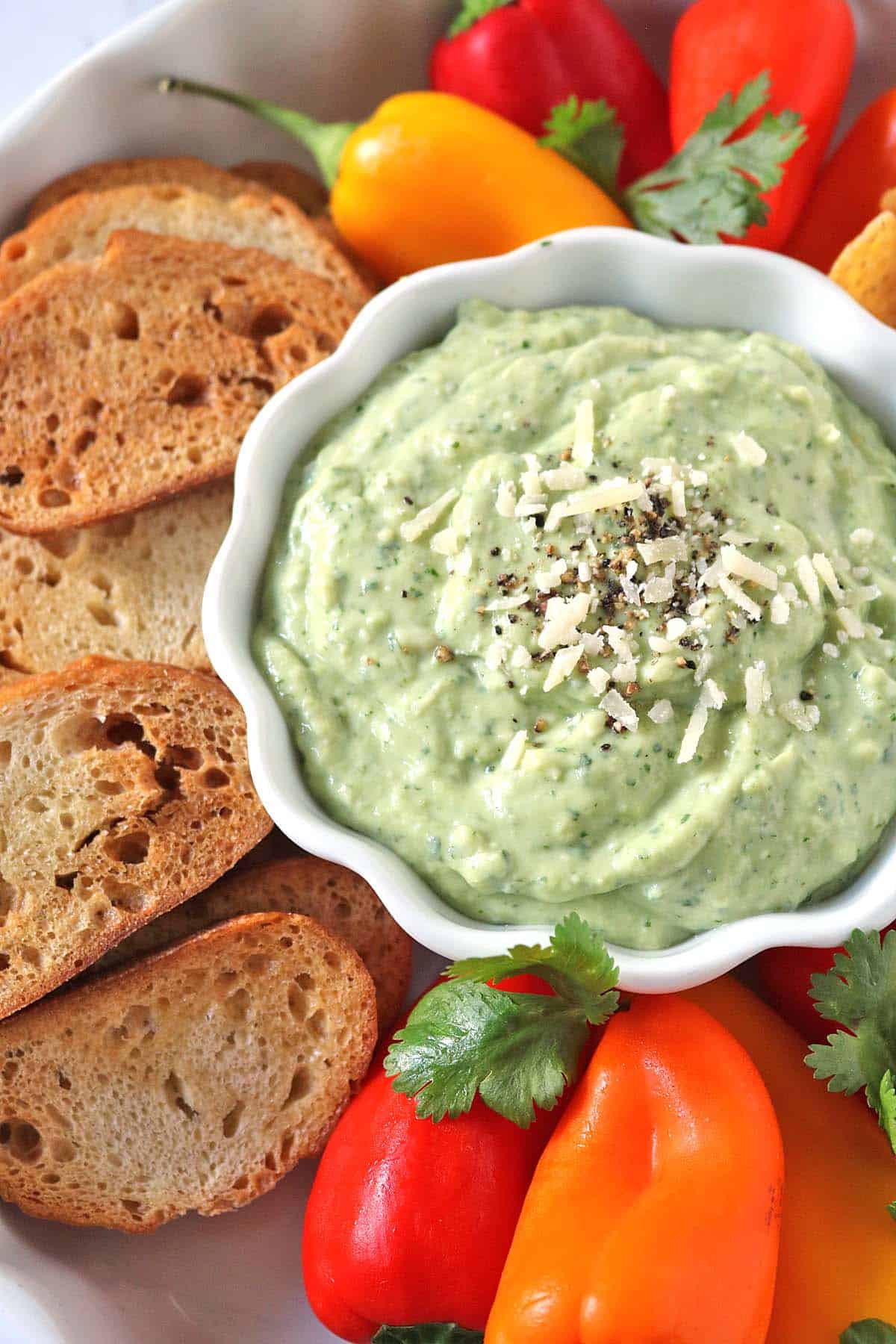 Creamy green dip topped with shredded cheese and pepper in a white bowl, surrounded by toasted bread slices, colorful mini bell peppers, and fresh cilantro.