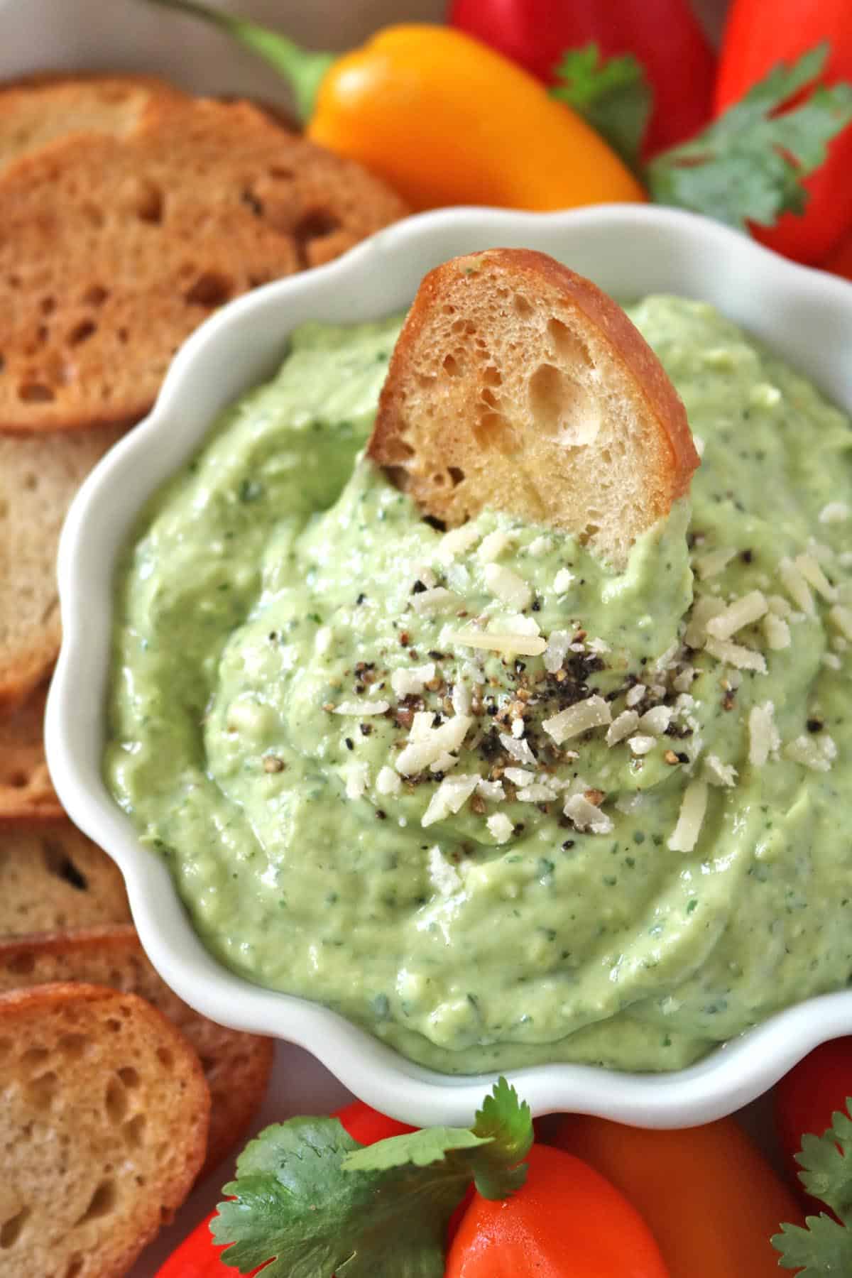 A bowl of creamy green dip topped with shredded cheese and cracked pepper, served with toasted bread slices and colorful mini bell peppers.