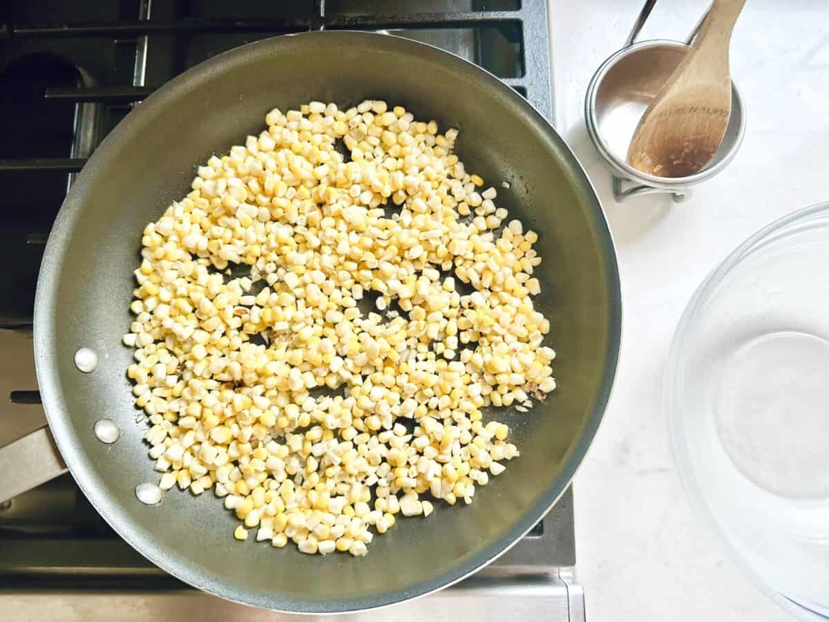 A frying pan with corn kernels on a stovetop, a small pot with milk and a wooden spoon, and an empty glass bowl nearby on a white countertop.