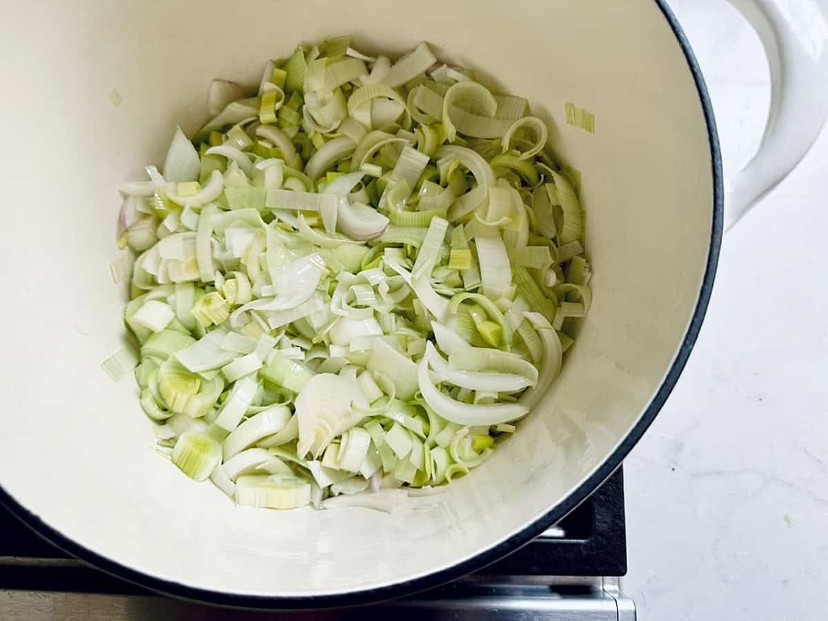 Chopped leeks in a white pot, ready to be cooked for a comforting Potato Leek Soup, sit on a stovetop with a light countertop in the background.