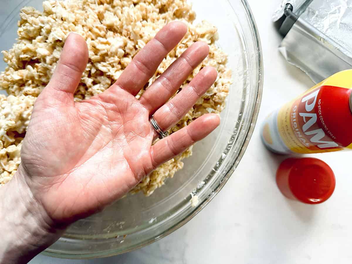 A persons open hand is held above a bowl of Rice Krispies mixture, with a can of PAM cooking spray and red cap on a white countertop nearby.