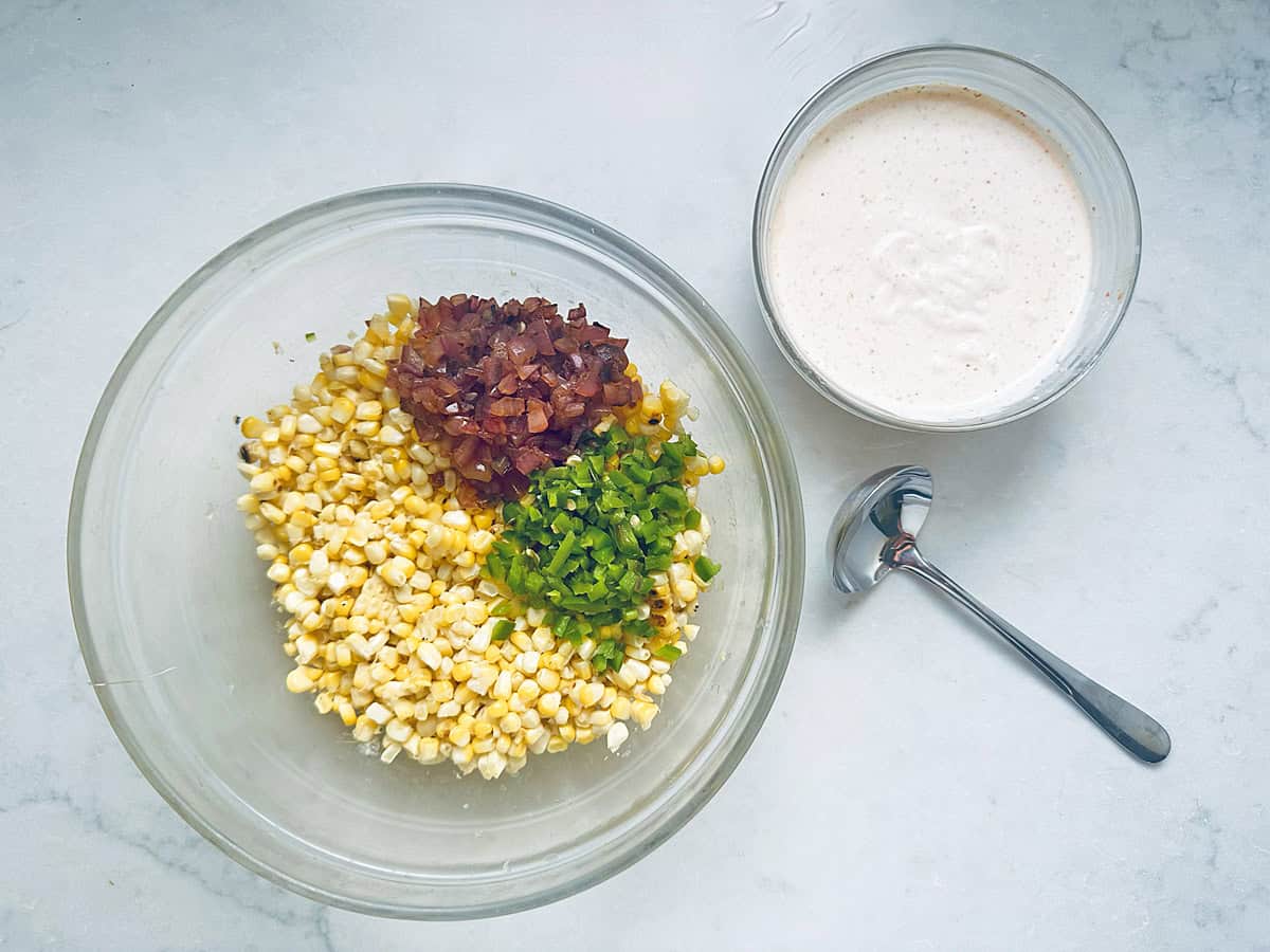 A glass bowl with chopped corn, red onion, and green pepper beside a bowl of creamy dressing and a spoon on a white surface—perfect fresh ingredients for assembling flavorful Grilled Street Corn Chicken Bowls.