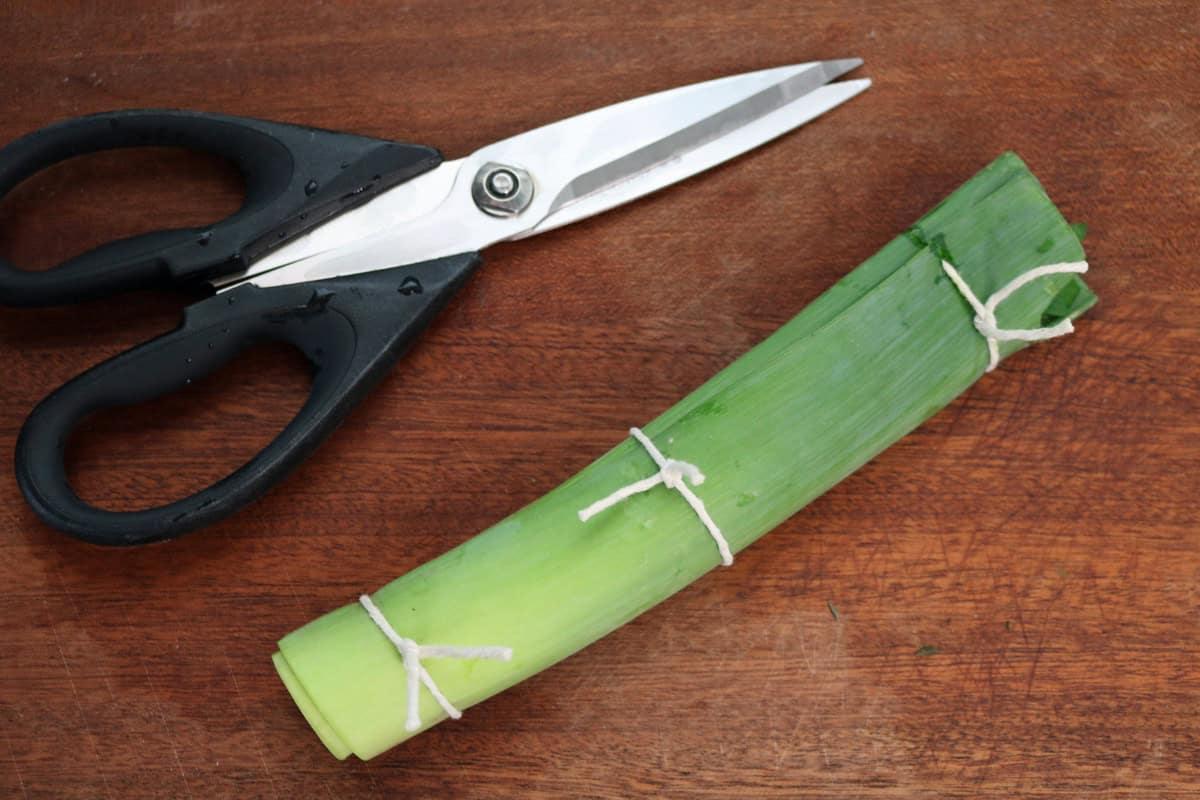A pair of black scissors next to a bundle of green leek stalks tied with white string, placed on a wooden surface—perfect for illustrating how to make a bouquet garni and sachet.