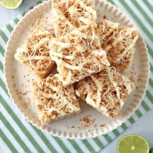 A plate of coconut-topped dessert bars sits on a white plate, surrounded by halved limes. The plate rests on a green-striped cloth on a white surface.