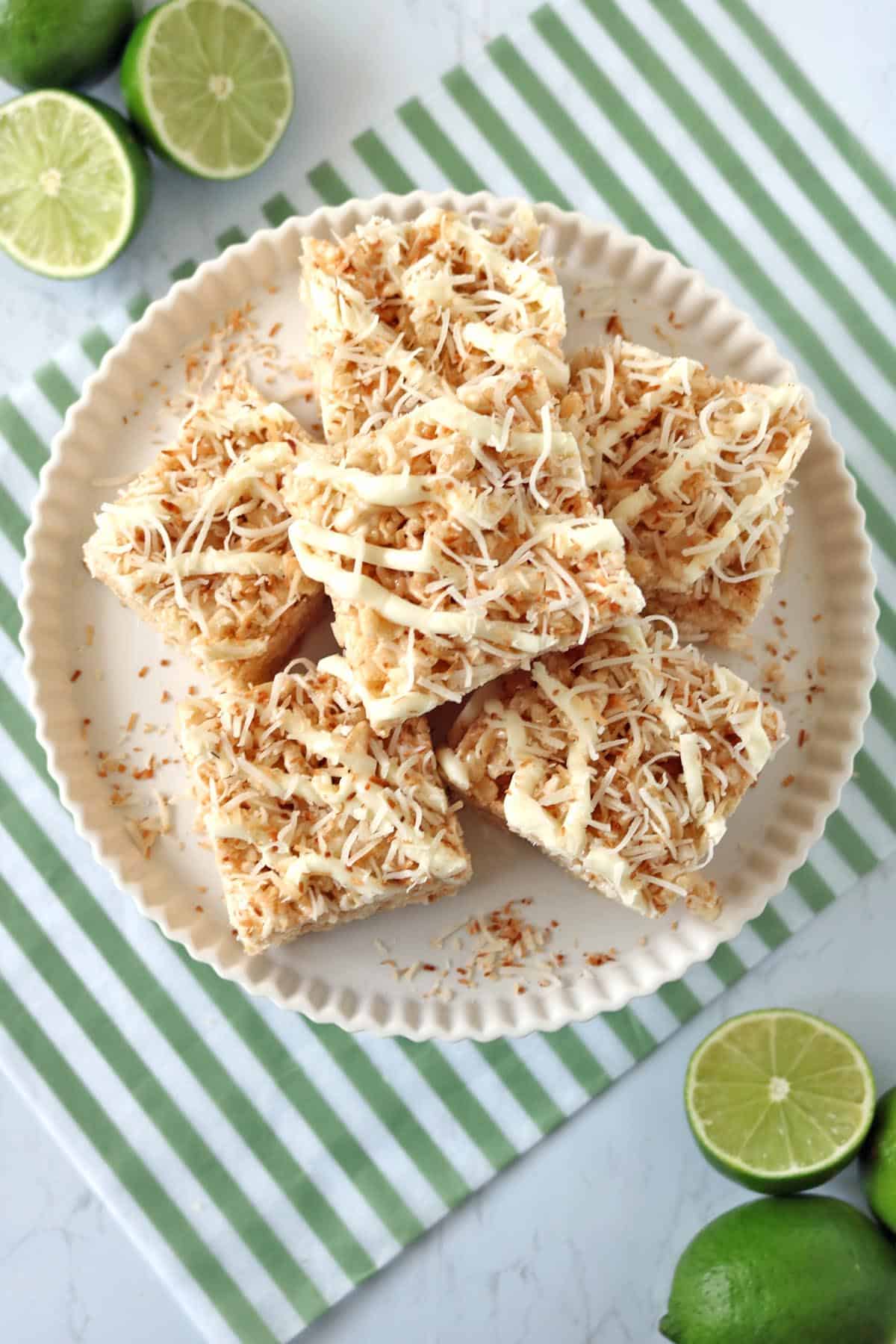 A plate of coconut-topped dessert bars sits on a white plate, surrounded by halved limes. The plate rests on a green-striped cloth on a white surface.