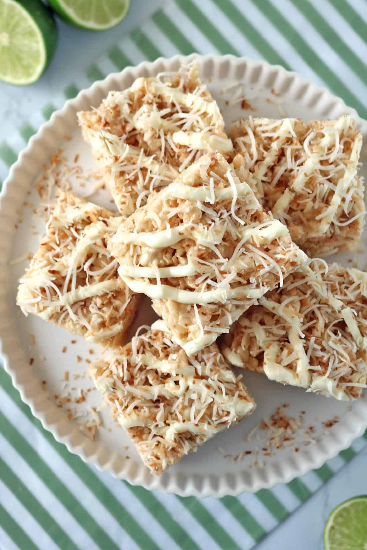 A plate of dessert bars topped with toasted coconut and white drizzle, arranged in a circle on a white plate. The plate sits on a green and white striped surface, with lime slices nearby.
