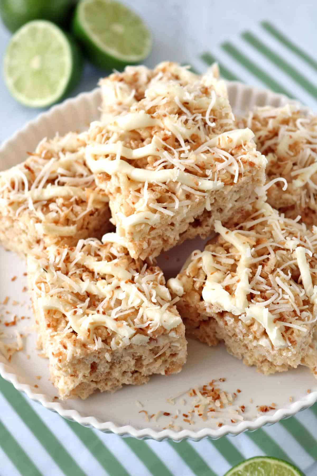 A plate of coconut Rice Krispies treats topped with toasted coconut and white chocolate drizzle, with sliced limes in the background on a green-striped cloth.