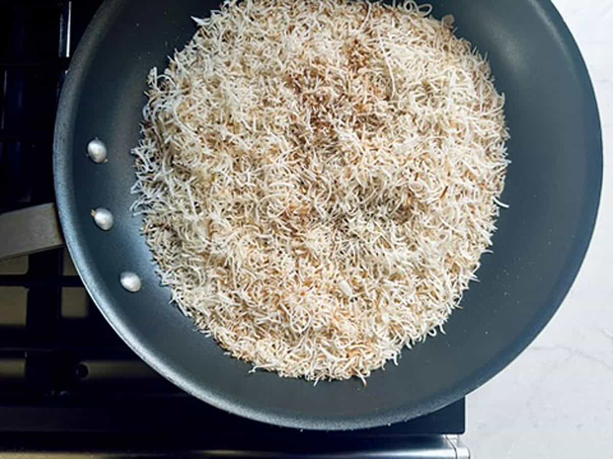 A nonstick pan on a stove contains lightly browned vermicelli noodles, some strands golden while others remain pale, indicating they are being toasted.