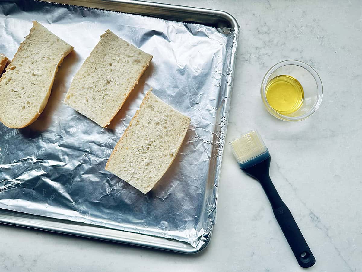 Three slices of bread on a foil-lined baking sheet, ready to be transformed into delicious Bacon Fig and Arugula Sandwiches, with a small bowl of olive oil and a basting brush beside them on a white countertop.