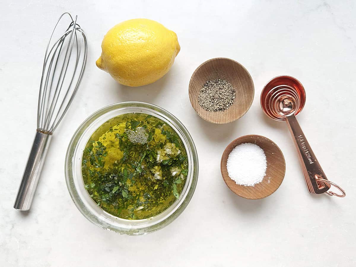 A whisk, lemon, bowl of herb vinaigrette, wooden bowls of salt and pepper, and copper measuring spoons arranged on a white surface—perfect essentials for preparing Foolproof French Grilled Chicken.