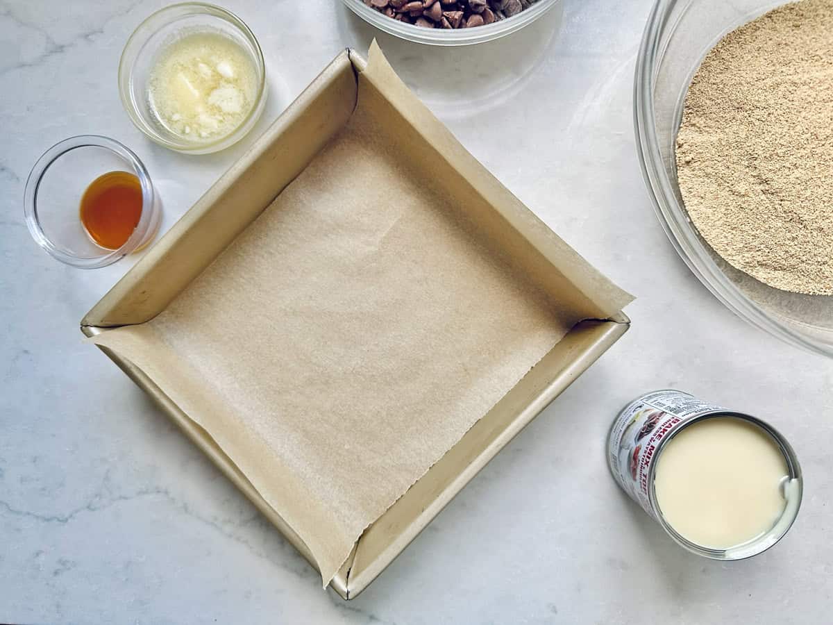 A square baking pan lined with parchment paper sits on a counter, ready for 1-2-3 Snap Brownies, surrounded by bowls of melted butter, vanilla extract, chocolate chips, graham cracker crumbs, and an open can of sweetened condensed milk.