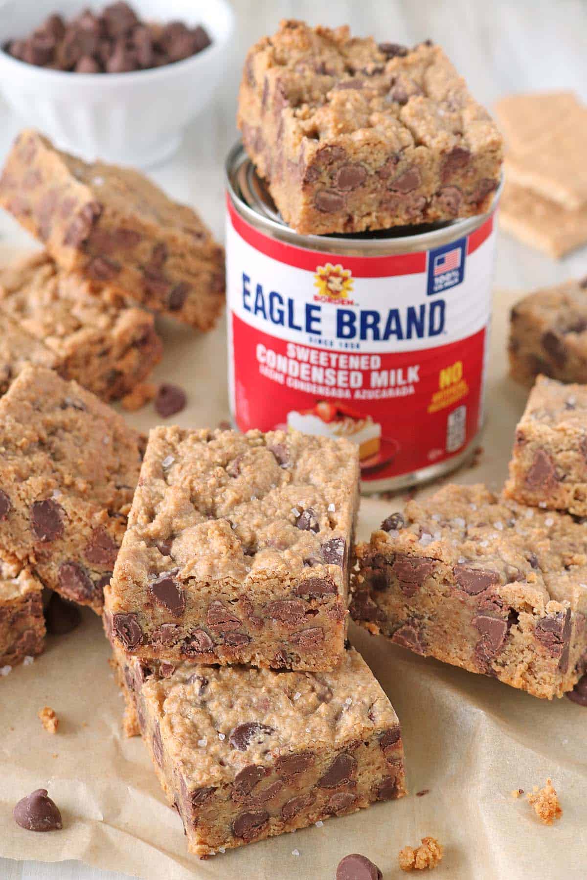 A can of Eagle Brand sweetened condensed milk sits surrounded by thick, chewy chocolate chip cookie bars sprinkled with sea salt. Some bars are stacked, reminiscent of irresistible 1-2-3 Snap Brownies, and a bowl of chocolate chips is visible in the upper left background on parchment paper.