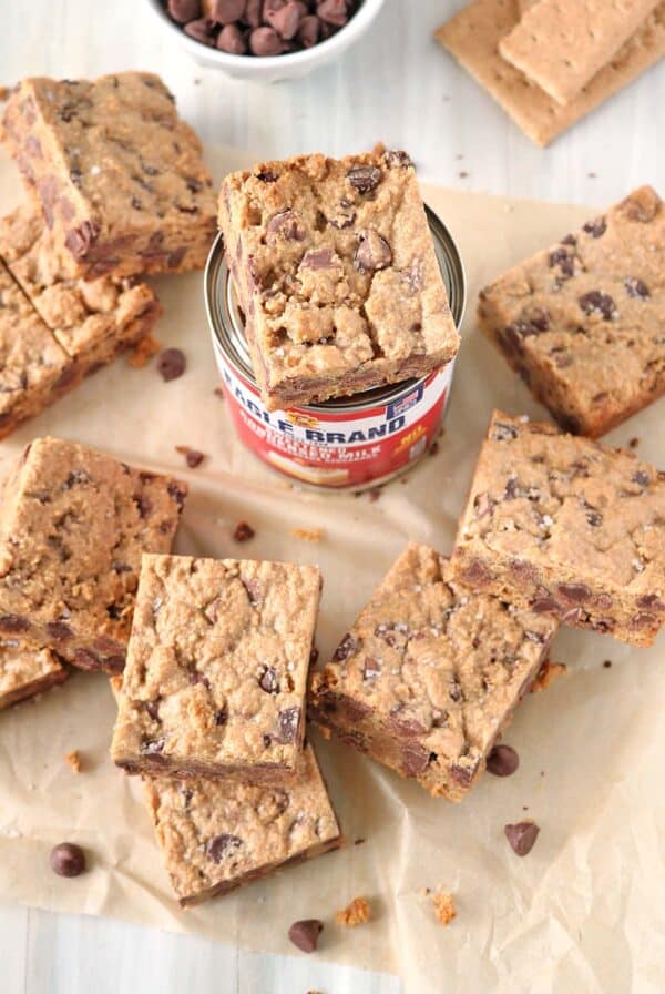 Top-down view of square 1-2-3 Snap Brownies scattered on parchment paper around a can of Eagle Brand sweetened condensed milk. A small bowl of chocolate chips and graham crackers are visible in the background.
