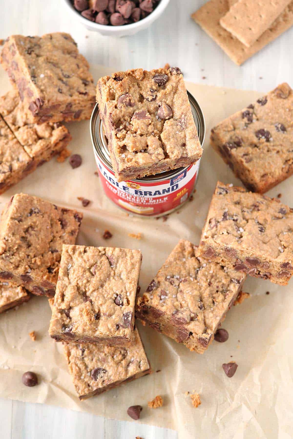 Top-down view of square 1-2-3 Snap Brownies scattered on parchment paper around a can of Eagle Brand sweetened condensed milk. A small bowl of chocolate chips and graham crackers are visible in the background.