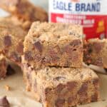 Two thick 1-2-3 Snap Brownies, packed with chocolate chips, are stacked on parchment paper, with a can of sweetened condensed milk and more bars in the background. The brownies have visible chocolate chips and a slightly crumbly texture.