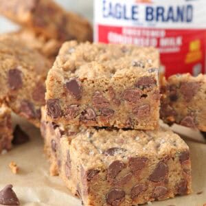 Two thick 1-2-3 Snap Brownies, packed with chocolate chips, are stacked on parchment paper, with a can of sweetened condensed milk and more bars in the background. The brownies have visible chocolate chips and a slightly crumbly texture.