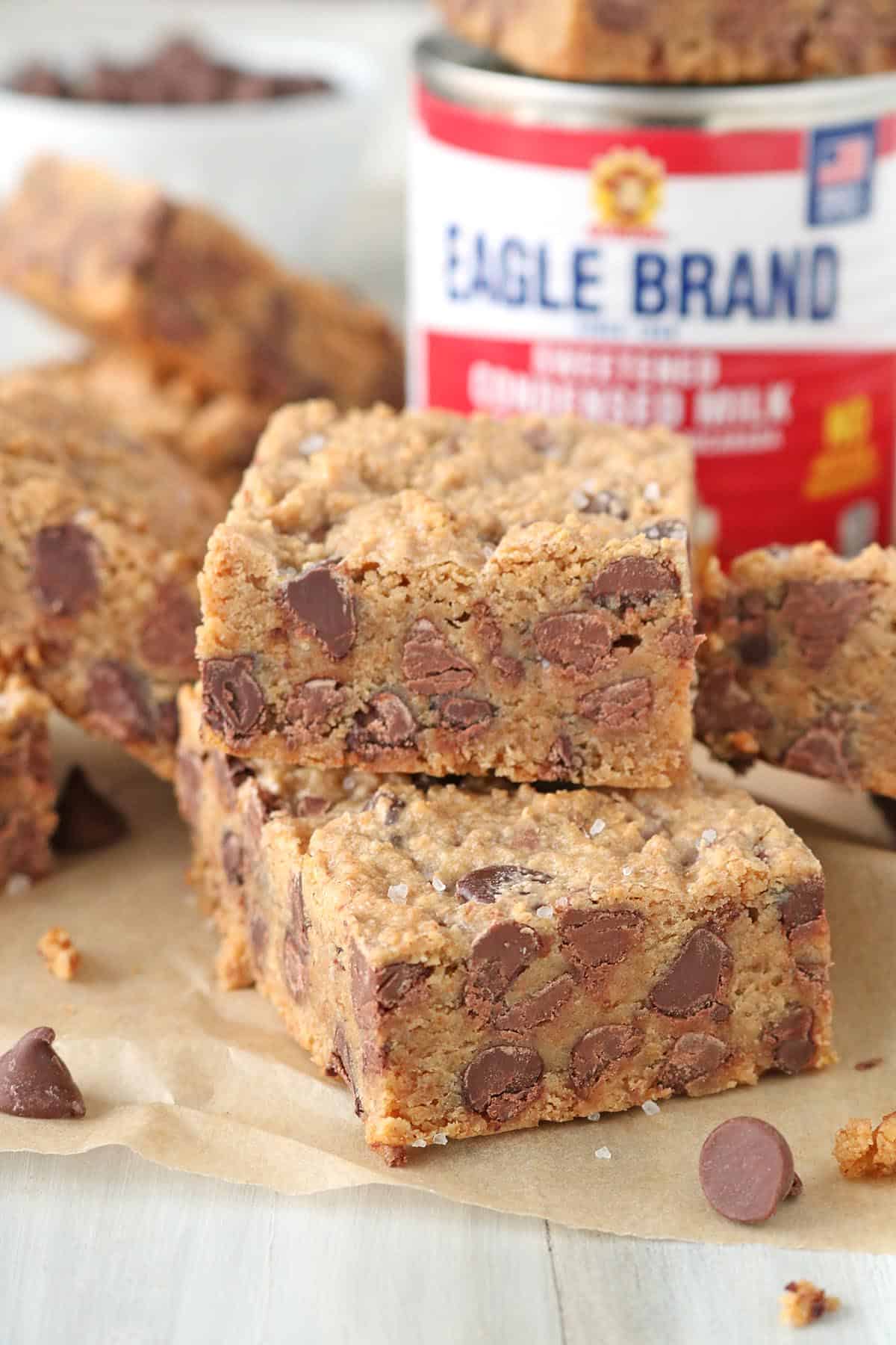 Two thick 1-2-3 Snap Brownies, packed with chocolate chips, are stacked on parchment paper, with a can of sweetened condensed milk and more bars in the background. The brownies have visible chocolate chips and a slightly crumbly texture.