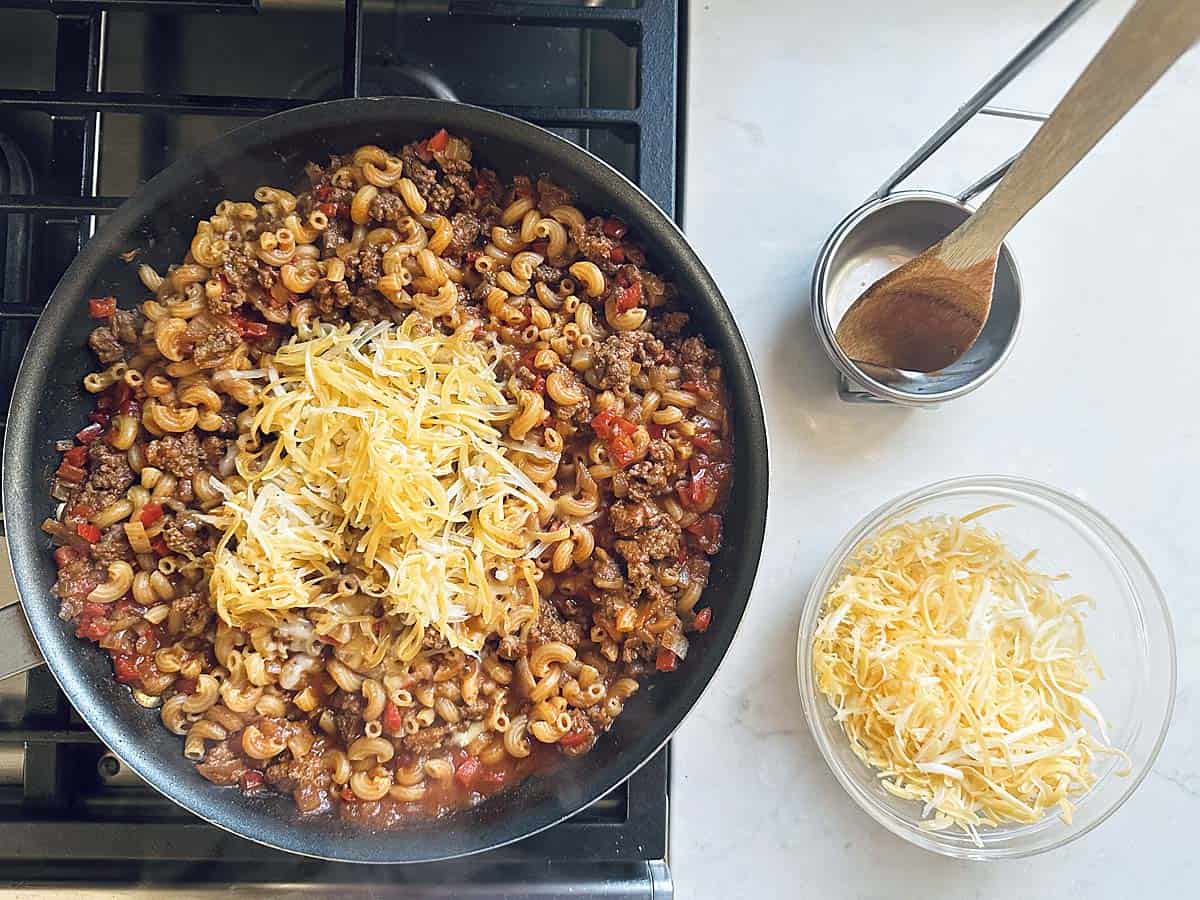 A skillet on a stove contains Skillet Cheesy Chili Mac with macaroni, ground beef, tomatoes, and shredded cheese. Nearby are a bowl of cheese, a measuring cup, and a wooden spoon on a white countertop.