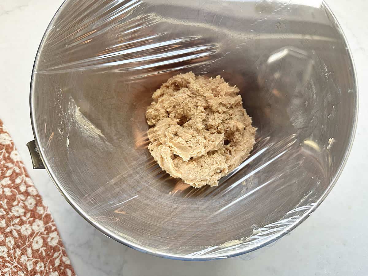 A metal mixing bowl with Apple Cider Snickerdoodles cookie dough covered in plastic wrap, sitting on a light countertop next to an orange patterned cloth.