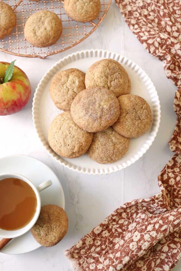 Plate of round Apple Cider Snickerdoodles on a white dish, with an apple, a cup of tea, and a cookie on a saucer nearby. Brown patterned cloth and a cooling rack with more cookies in the background.