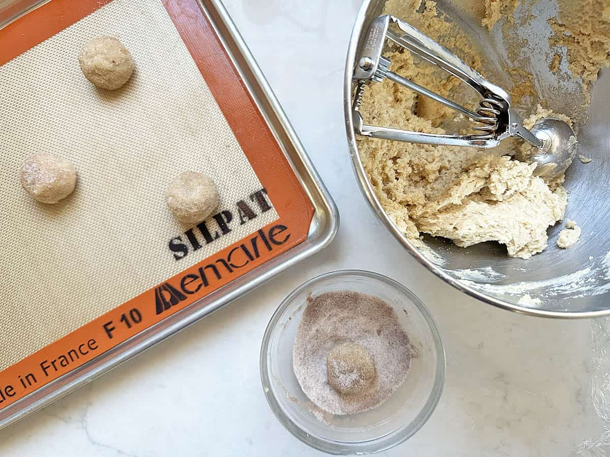 Apple Cider Snickerdoodles cookie dough balls being rolled in cinnamon sugar, placed on a baking sheet lined with a Silpat mat, with a bowl of dough and a cookie scoop nearby.