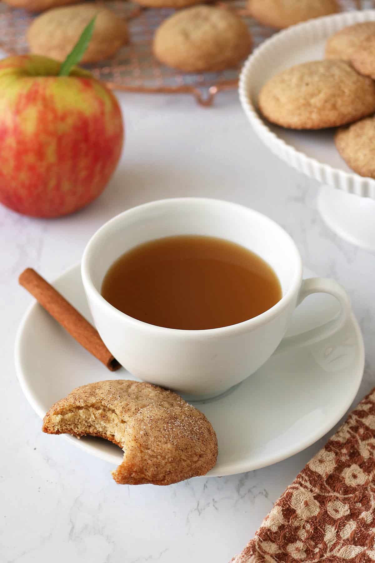 A white cup of tea sits on a saucer with a cinnamon stick and a bitten cookie. An apple, Apple Cider Snickerdoodles on a stand, and cookies on a cooling rack are in the background.