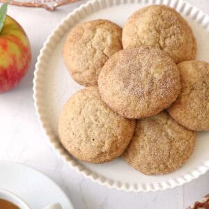 A white plate with round, cinnamon sugar Apple Cider Snickerdoodles on a marble surface, next to a red apple, a cup of tea, and a cooling rack with more cookies in the background.