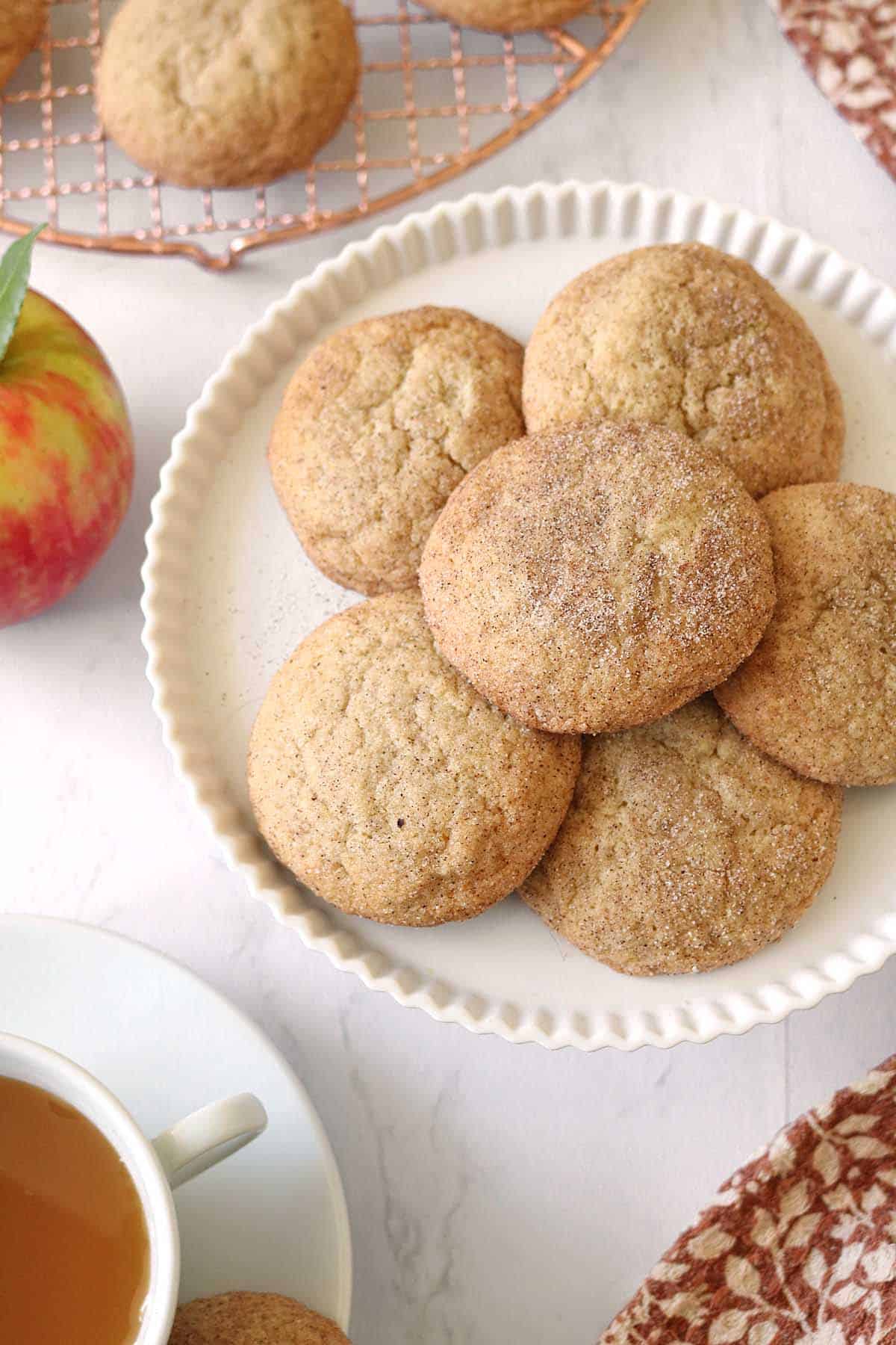 A white plate with round, cinnamon sugar Apple Cider Snickerdoodles on a marble surface, next to a red apple, a cup of tea, and a cooling rack with more cookies in the background.
