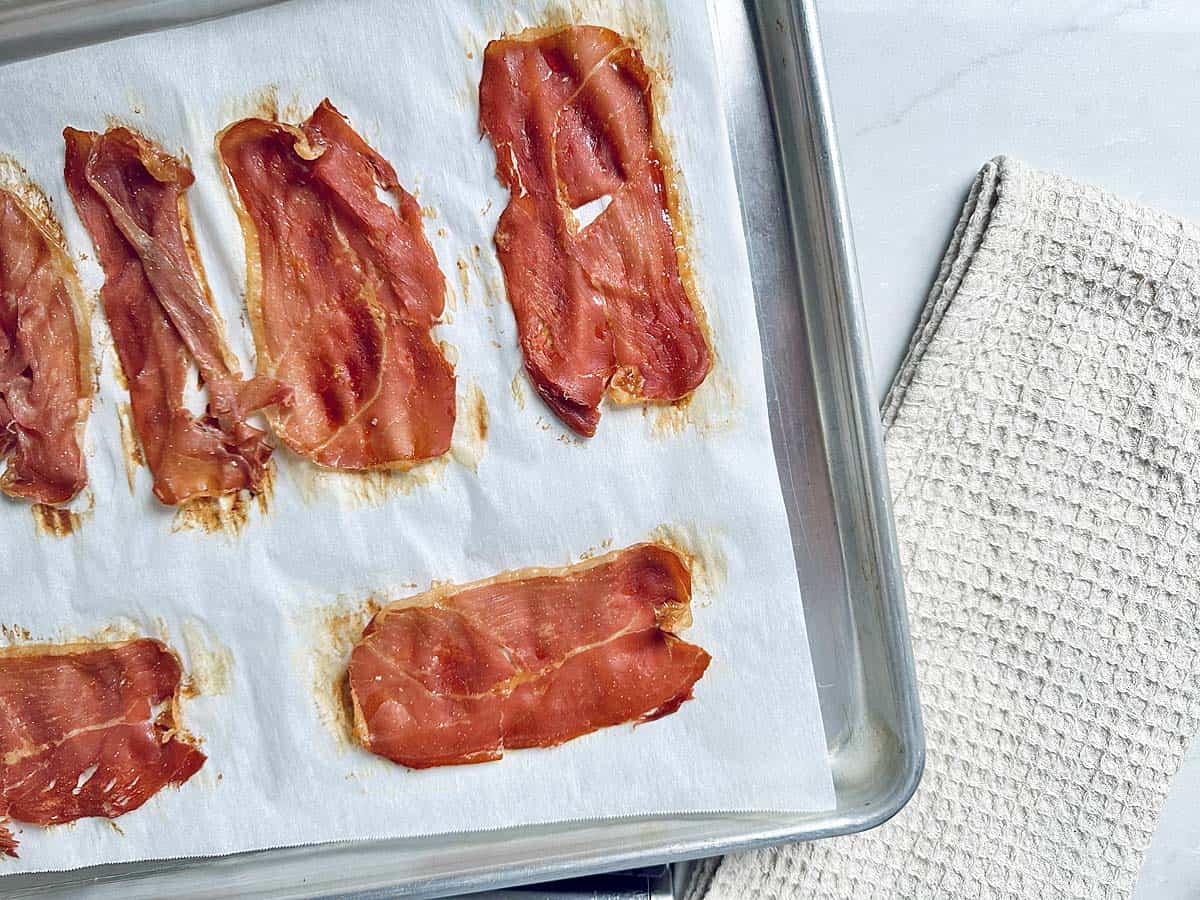 Slices of prosciutto on parchment paper atop a baking sheet, next to a beige textured kitchen towel, on a white surface—perfect as a savory topping for your Kale Apple Harvest Salad.