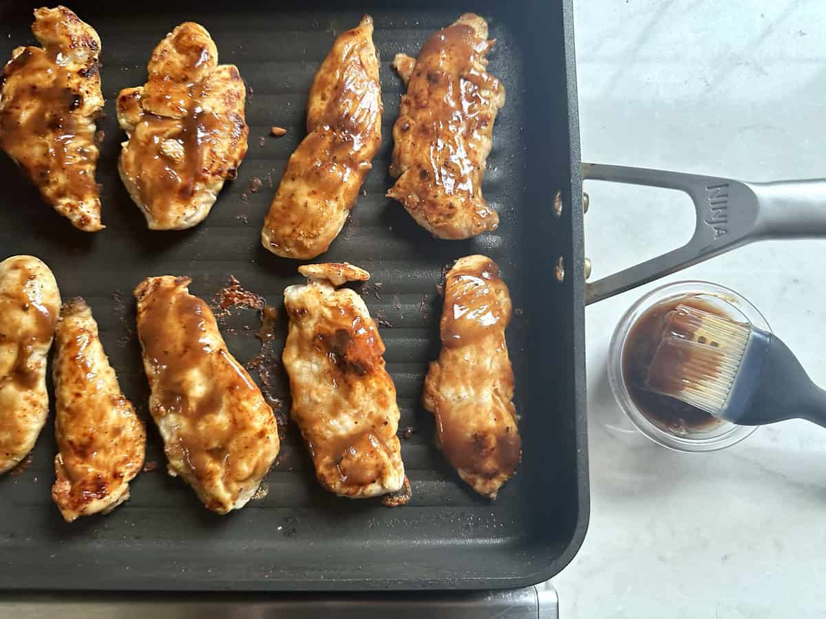 Chicken strips cooking on a grill pan, glazed with sauce. A small cup with sauce and a basting brush sits beside the pan on a light kitchen countertop.