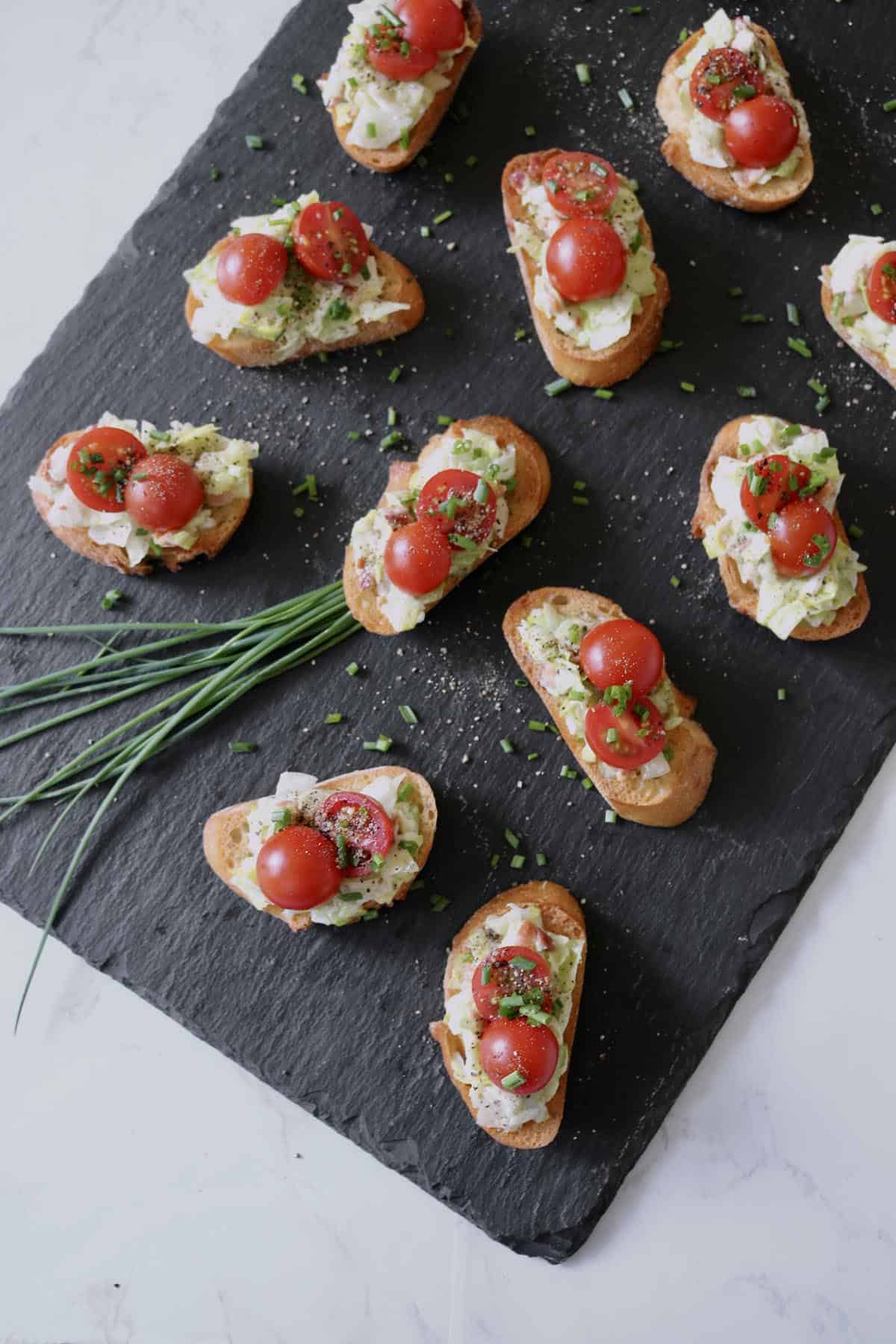 Slices of toasted bread topped with creamy cheese, halved cherry tomatoes, and chopped herbs are arranged on a black slate serving board with a bunch of fresh chives beside them.