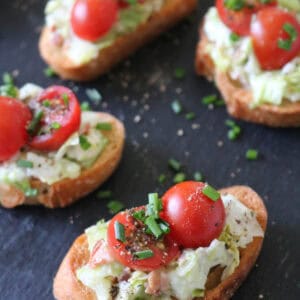 Five crostini topped with lettuce, creamy spread, halved cherry tomatoes, chopped chives, and black pepper sit on a dark slate surface.