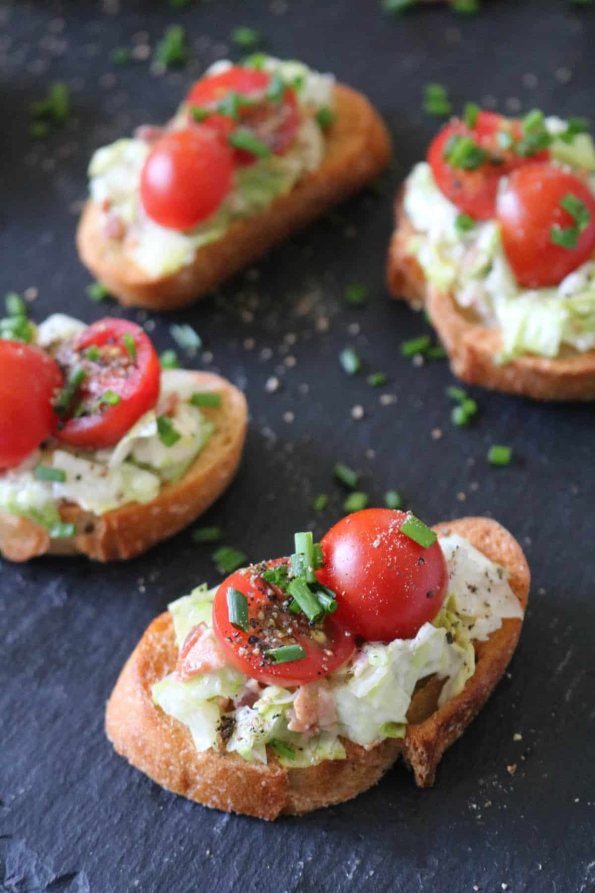 Five crostini topped with lettuce, creamy spread, halved cherry tomatoes, chopped chives, and black pepper sit on a dark slate surface.