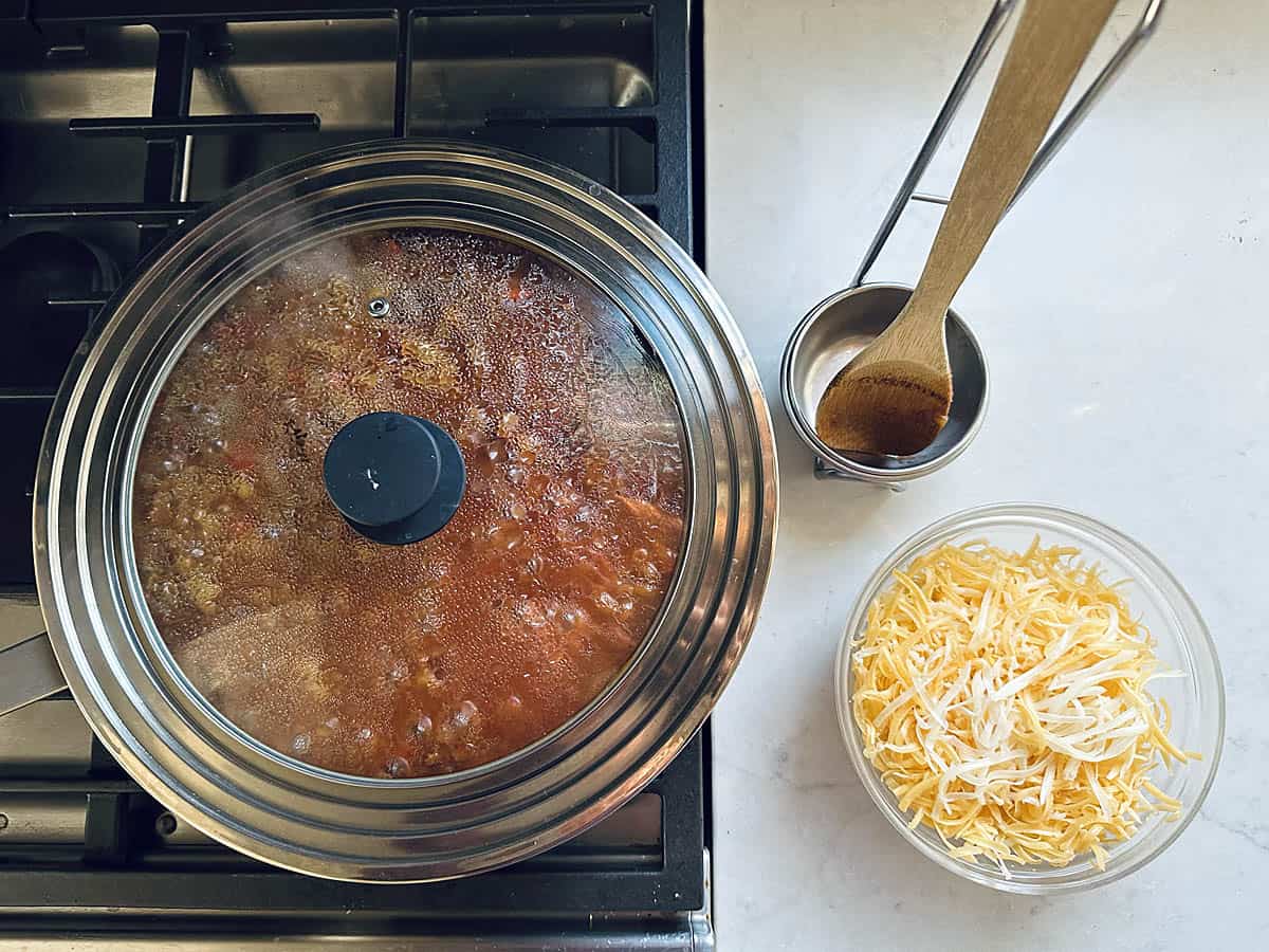 A pot with a glass lid simmers on a stove. Beside it, there’s a bowl of shredded cheese—perfect for making Skillet Cheesy Chili Mac—and a metal cup holding a wooden spoon with some spices or sauce on it.