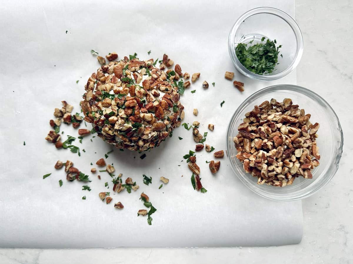 Vivian Howard’s Party Magnet cheese ball coated with chopped pecans and herbs rests on parchment paper, with small bowls of chopped parsley and pecans nearby, all arranged on a marble surface.