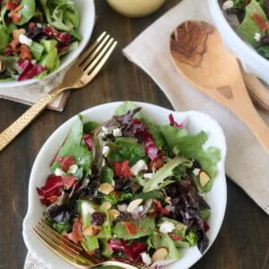 A bowl of mixed green salad with sliced almonds, crumbled cheese, and bacon bits sits on a wooden table, evoking the signature style of The Galley Gourmet. A gold fork and dressing add elegant touches in the background.