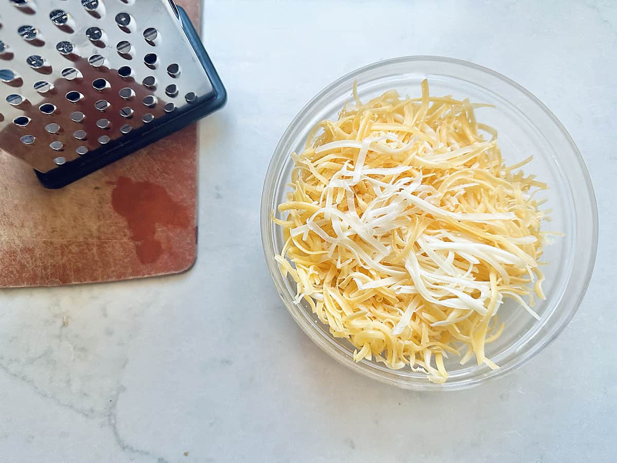 A glass bowl filled with shredded cheese sits on a white surface next to a metal box grater on a small wooden cutting board, ready to top your Skillet Cheesy Chili Mac.