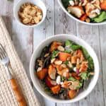 Bowl of salad with grilled chicken, peaches, greens, red onions, feta, and sliced almonds on a wooden table, with a small bowl of almonds and a fork nearby.