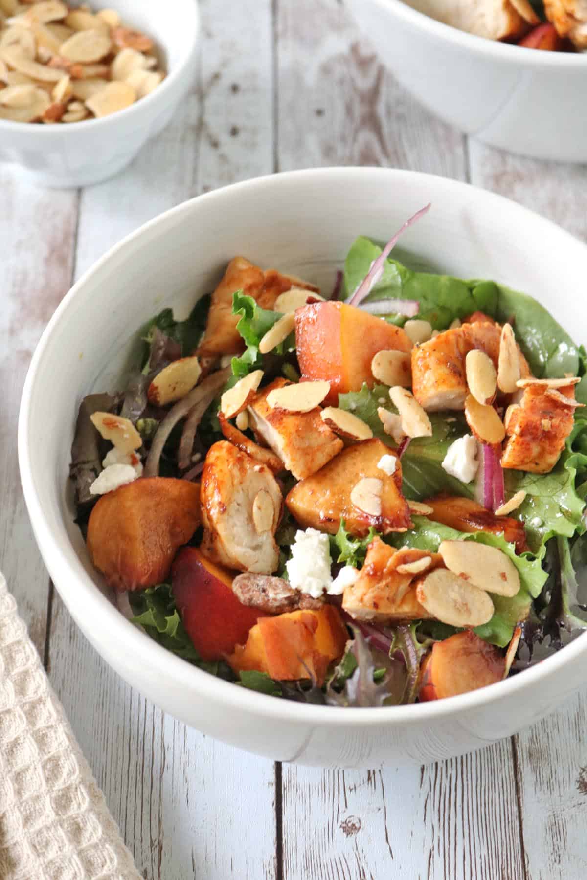 A white bowl filled with salad greens, grilled chicken pieces, peach slices, red onions, crumbled cheese, and toasted almond slices, set on a rustic wooden table.