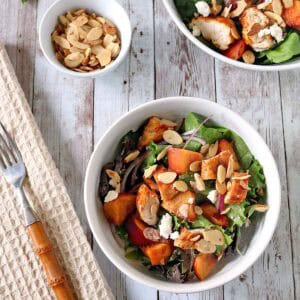 Bowl of salad with grilled chicken, peaches, greens, red onions, feta, and sliced almonds on a wooden table, with a small bowl of almonds and a fork nearby.