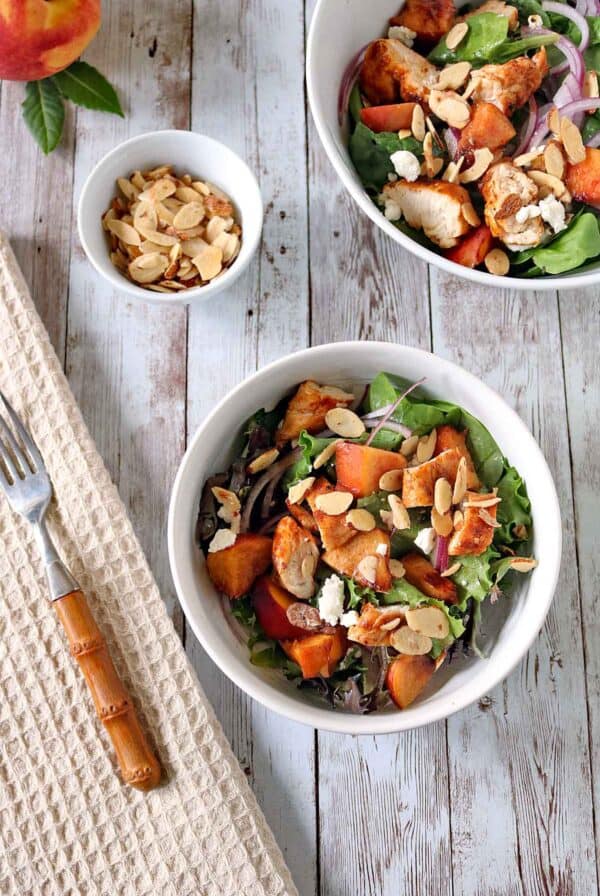 Bowl of salad with grilled chicken, peaches, greens, red onions, feta, and sliced almonds on a wooden table, with a small bowl of almonds and a fork nearby.