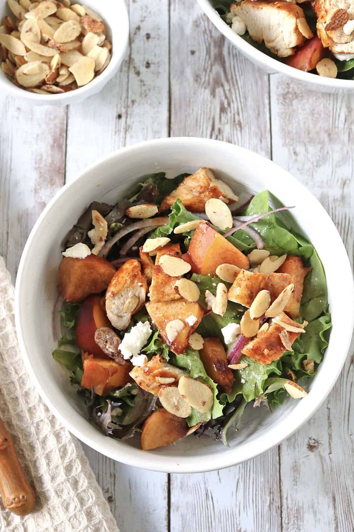 A white bowl filled with mixed greens, grilled chicken pieces, peach slices, crumbled cheese, and toasted almond slivers, on a rustic wooden table.