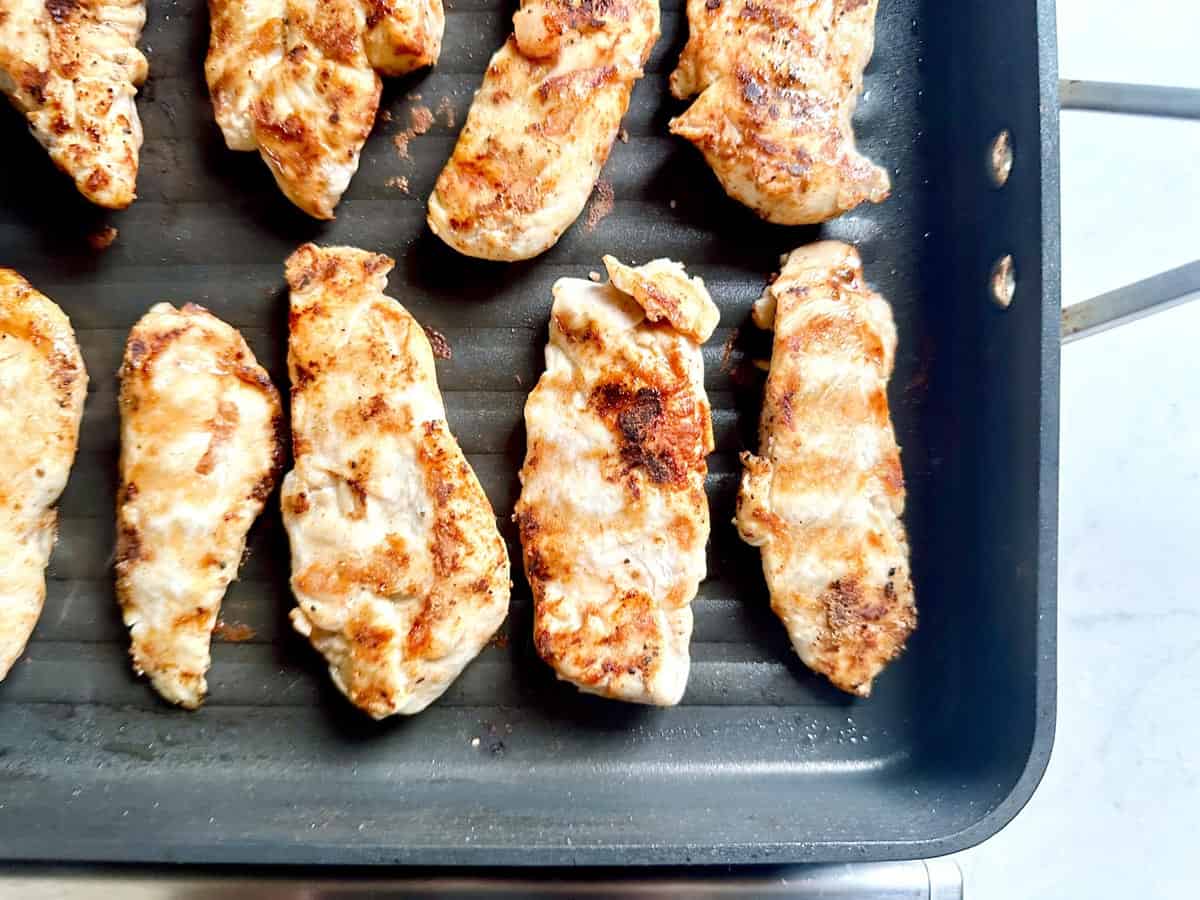 Grilled chicken strips cooking on a ridged grill pan, showing golden brown sear marks. The pan is black with metal handles, and the background is a light kitchen surface.
